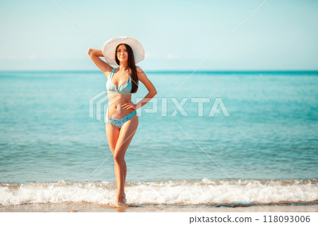 A beautiful woman in a bikini and a beach hat poses standing on the beach. In the background, the sea and sky. Copy space. The concept of beach holidays and summer vacation 118093006