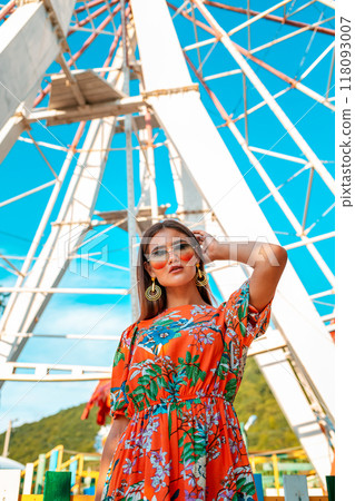 A beautiful woman in a dress and glasses, posing against the background of a damn wheel in an amusement Park. Bottom view. The concept of fashion and entertainment 118093007