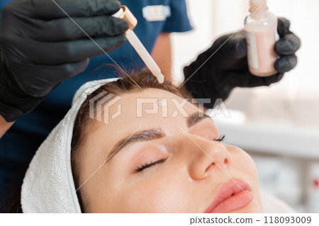 A cosmetologist drips a cosmetic serum on a woman's forehead during the beauty procedure. Close-up portrait of the client. Professional skin care in salon A cosmetologist drips a cosmetic serum on a woman's forehead during the beauty procedure. Close-up portrait of the client. Professional skin care in salon 118093009
