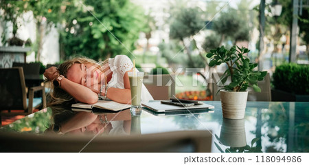 Woman resting her head on a notebook at a cafe amid lush greenery in the afternoon 118094986