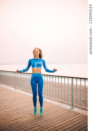 Young woman wearing blue workout attire skipping on a boardwalk by the seaside during sunrise 118095014