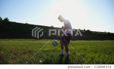 Sportsman juggling soccer ball on stadium at sunny day. Young man kicking ball at green field. Professional footballer practicing tricks at meadow with sunlight at background. Freestyle football. 118096358