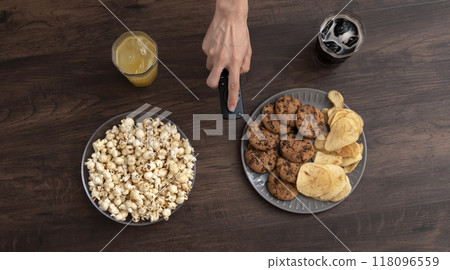 一個人在擺著零食和糖果的桌子上操作遙控器。打開電視，觀看視訊串流，觀看錄音 118096559