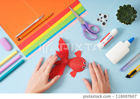 Top view of paper cutting tools, scissors, cutter, female hands holding finished red fish paper crafts. DIY project concept for kids, hobbies and creativity. Top view of paper cutting tools, scissors, cutter, female hands holding finished red fish paper crafts. DIY project concept for kids, hobbies and creativity. 118097607