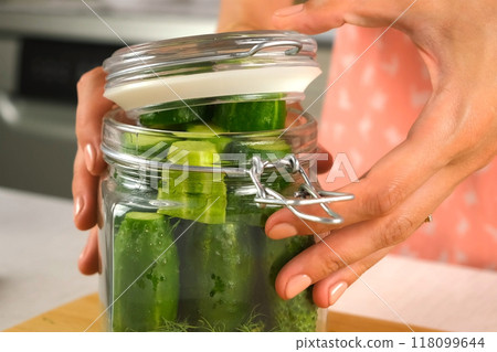 Woman puts cucumbers in glass jar to cook pickles closing lid, hands close-up. 118099644