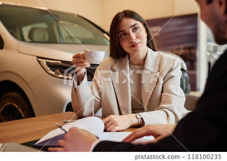 Successful young woman drinking coffee at table while signing purchase contract buying new car at dealership center 118100235