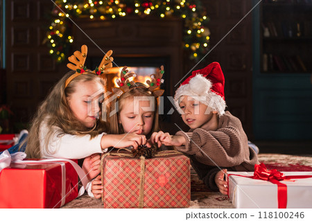 Merry Christmas Xmas holiday. Children unpacking open up gift box near Christmas tree. Kids in living room with Christmas tree fireplace opening gift box with surprise faces. Christmas eve at home Merry Christmas Xmas holiday. Children unpacking open up gift box near Christmas tree. Kids in living room with Christmas tree fireplace opening gift box with surprise faces. Christmas eve at home 118100246