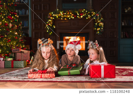 Merry Christmas Xmas holiday. Children unpacking open up gift box near Christmas tree. Kids in living room with Christmas tree fireplace opening gift box with surprise faces. Christmas eve at home Merry Christmas Xmas holiday. Children unpacking open up gift box near Christmas tree. Kids in living room with Christmas tree fireplace opening gift box with surprise faces. Christmas eve at home 118100248