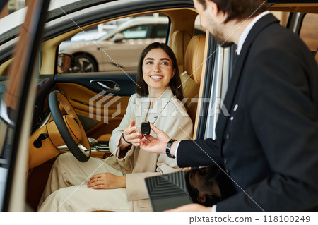 Satisfied female client at drivers seat of premium car with beige leather cabin taking key from agent, while buying or renting vehicle at car rental office or dealership center 118100249
