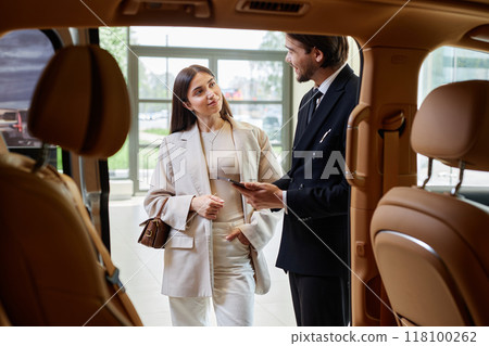 Shot from car cabin with beige leather interior of smiling well dressed woman consulting with auto dealer on buying new vehicle at dealership center 118100262