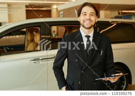 Portrait of smiling adult Central Asian male auto dealer in formal black suit holding tablet and looking at camera posing at car dealership center, copy space Portrait of smiling adult Central Asian male auto dealer in formal black suit holding tablet and looking at camera posing at car dealership center, copy space 118100276