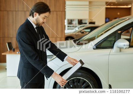 Side view of Central Asian male auto dealer in black suit thoroughly reading information on car dealership stand displaying vehicle spec sheets at dealership center, copy space 118100277