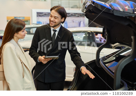 Medium shot of smiling Central Asian auto dealer in black suit advising female customer on new car while demonstrating trunk capacity at dealership center 118100289
