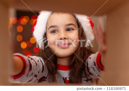 Happy girl in New Year's pajamas and Santa Claus hat takes out a gift Happy girl in New Year's pajamas and Santa Claus hat takes out a gift 118100971