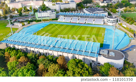 A high-quality aerial view of a modern sports stadium, featuring a football field and running track, surrounded by lush greenery and urban landscape. A high-quality aerial view of a modern sports stadium, featuring a football field and running track, surrounded by lush greenery and urban landscape. 118101350