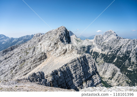 The majestic Velika Mojstrovka and Mangart mountains rise dramatically in the Julian Alps, showcasing their rugged beauty under a clear blue sky. A perfect destination for hikers and nature lovers. 118101396