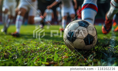 A close-up shot of a soccer ball being kicked during a lively match, with players in uniform running on a green field 118102216