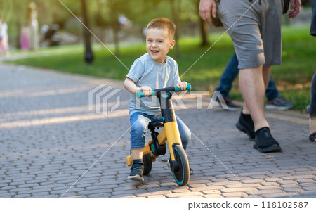 A cute smiling toddler boy of three or four years old rides a bicycle or balance bike in a city park on a sunny summer day. Toddlerhood and childhood concept. Selective focus A cute smiling toddler boy of three or four years old rides a bicycle or balance bike in a city park on a sunny summer day. Toddlerhood and childhood concept. Selective focus 118102587