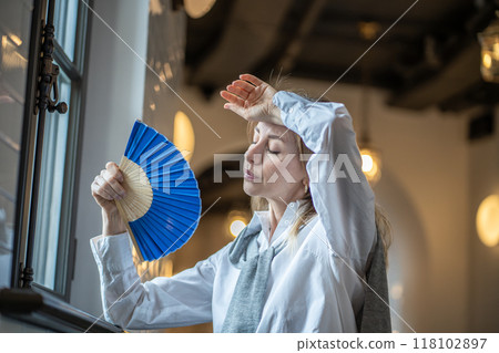 Tired and dissatisfied woman fans herself and feels fainting in stuffy hot office in summer heat 118102897