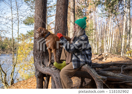 Serenity woman petting dog, sits on tree roots in autumn forest enjoys tranquility time on nature. 118102902