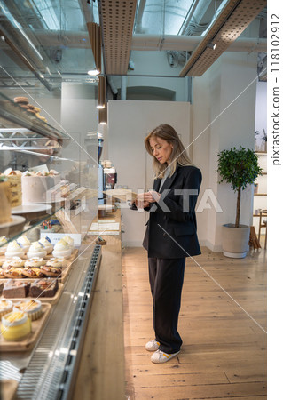 Serious businesswoman in black suit reading menu, standing at showcase with desserts in bakery cafe. 118102912