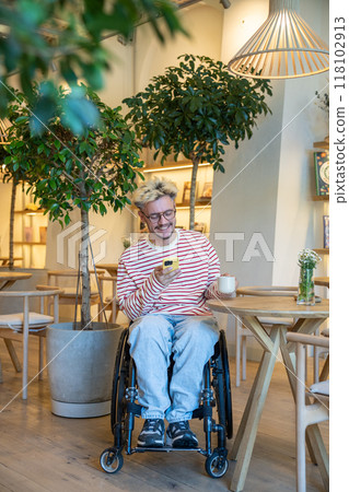 Happy, laughing disabled man in wheelchair chats in social media on smartphone on coffee break 118102913