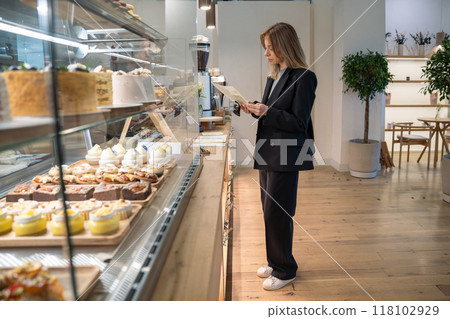 Focused interested woman in bakery choosing sweets pastries, browsing menu for holiday desserts. Focused interested woman in bakery choosing sweets pastries, browsing menu for holiday desserts. 118102929