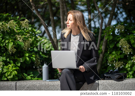 Interested woman with laptop on knees working remotely online on fresh air near office on sunny day. 118103002