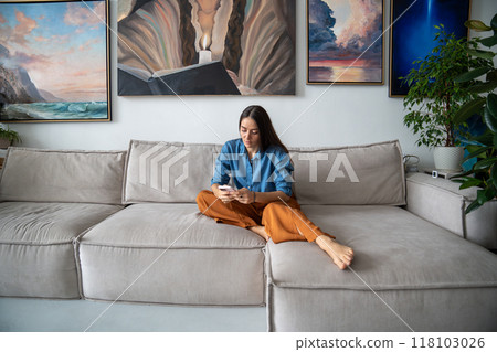 Relaxed woman holding phone chatting, checking news sits on couch in creative apartment with plants 118103026