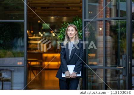 Thoughtful confident woman with laptop in hands standing at entrance of cafe in morning pondering. 118103028