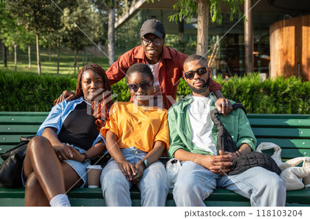 Group of happy african american students sits on bench in university campus looks at camera together Group of happy african american students sits on bench in university campus looks at camera together 118103204