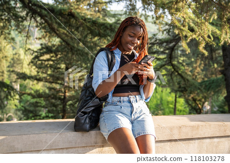 African American young woman smiling, engrossed in smartphone, chats with friends in sunny park 118103278