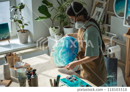 Craftswoman in respirator inspect dried epoxy resin painting with waves, checking result in workshop Craftswoman in respirator inspect dried epoxy resin painting with waves, checking result in workshop 118103318