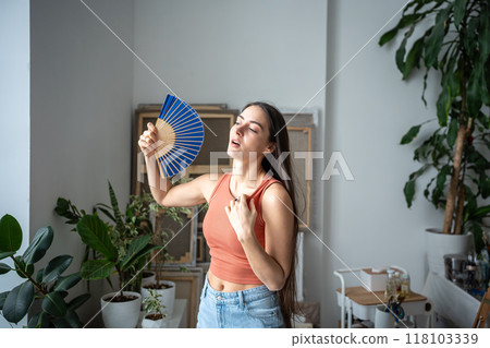 Tired woman cooling herself with paper fan at home, suffering from heat and lack of air conditioning Tired woman cooling herself with paper fan at home, suffering from heat and lack of air conditioning 118103339