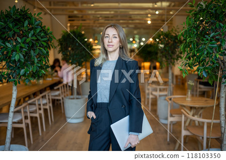 Businesslike confident woman in suit with laptop in hand standing in hall restaurant looks at camera 118103350