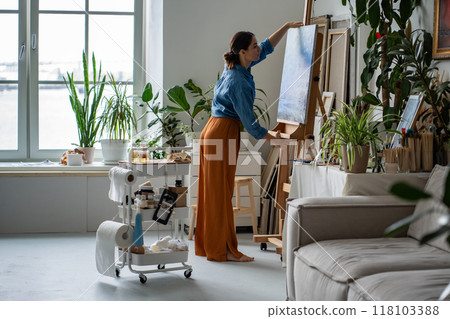 Craftswoman mounting finalized blue painting on easel, taking canvas in hands in radiant art gallery 118103388
