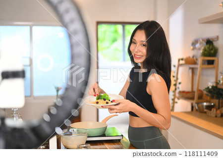 Attractive young female blogger recording video vlog about preparing a nutritious recipe in kitchen 118111409