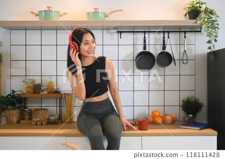 Relaxed young sports woman having fun listening to music in headphone, sitting on kitchen counter Relaxed young sports woman having fun listening to music in headphone, sitting on kitchen counter 118111428