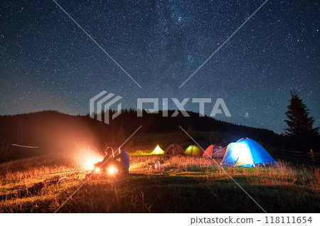 Night camping in mountains under starry sky. Silhouette of two people, couple sitting on grass near campsite, admiring landscape. Concept of tourism and healthy living. 118111654