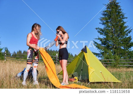 Two women hikers camping in mountains. Female tourists inflating the air mattress, wearing sportswear, hiking, making campsite. Concept of tourism and traveling. 118111658