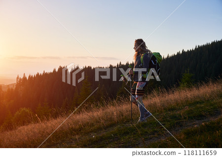 Woman hiker hiking outdoors at sunset. Sporty, positive woman traveling in mountains. Young, sporty female with backpack using trekking sticks, walking. Concept of harmony with nature. 118111659