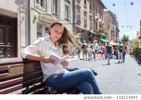 A young woman enjoys reading on a sunny day while seated on a bench in a lively street filled with pedestrians and vibrant shops A young woman enjoys reading on a sunny day while seated on a bench in a lively street filled with pedestrians and vibrant shops 118113845
