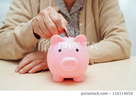 Retired elderly woman putting coins money in piggy bank and worry about monthly expenses and treatment fee payment. 118114058