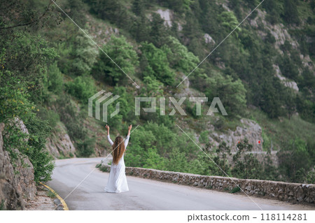 A woman is standing on a road, looking up at the sky. She is wearing a white dress and she is happy. The scene is set in a mountainous area, with trees and rocks in the background. A woman is standing on a road, looking up at the sky. She is wearing a white dress and she is happy. The scene is set in a mountainous area, with trees and rocks in the background. 118114281