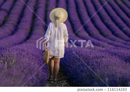 A woman wearing a straw hat walks through a field of purple flowers 118114283