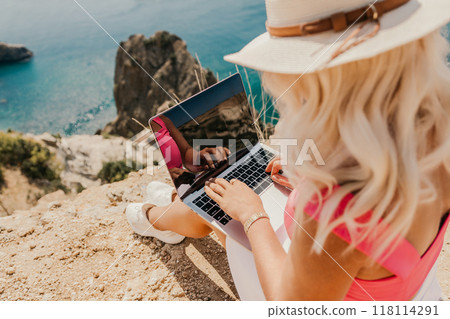 A woman is sitting on a rock by the ocean, typing on her laptop. Concept of relaxation and leisure, as the woman enjoys the beautiful view of the water while working on her computer. 118114291