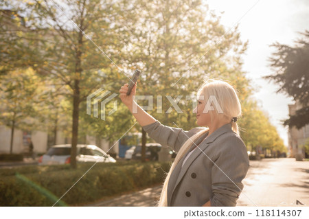 A woman is taking a picture of herself with her cell phone. She is wearing a gray jacket and scarf. The scene is set in a city with trees and cars in the background. 118114307