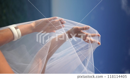 A woman is holding a white veil over her head. The woman is standing on a beach, and the ocean is visible in the background. A woman is holding a white veil over her head. The woman is standing on a beach, and the ocean is visible in the background. 118114405