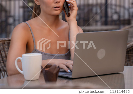 Woman Laptop Phone Call Cafe - A woman sits at a cafe table with a laptop and a phone, talking on the phone while working. Woman Laptop Phone Call Cafe - A woman sits at a cafe table with a laptop and a phone, talking on the phone while working. 118114455