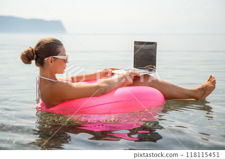 A woman is sitting in a pink inflatable raft on a lake, using a laptop. Concept of relaxation and leisure, as the woman is enjoying her time outdoors while working on her laptop. A woman is sitting in a pink inflatable raft on a lake, using a laptop. Concept of relaxation and leisure, as the woman is enjoying her time outdoors while working on her laptop. 118115451